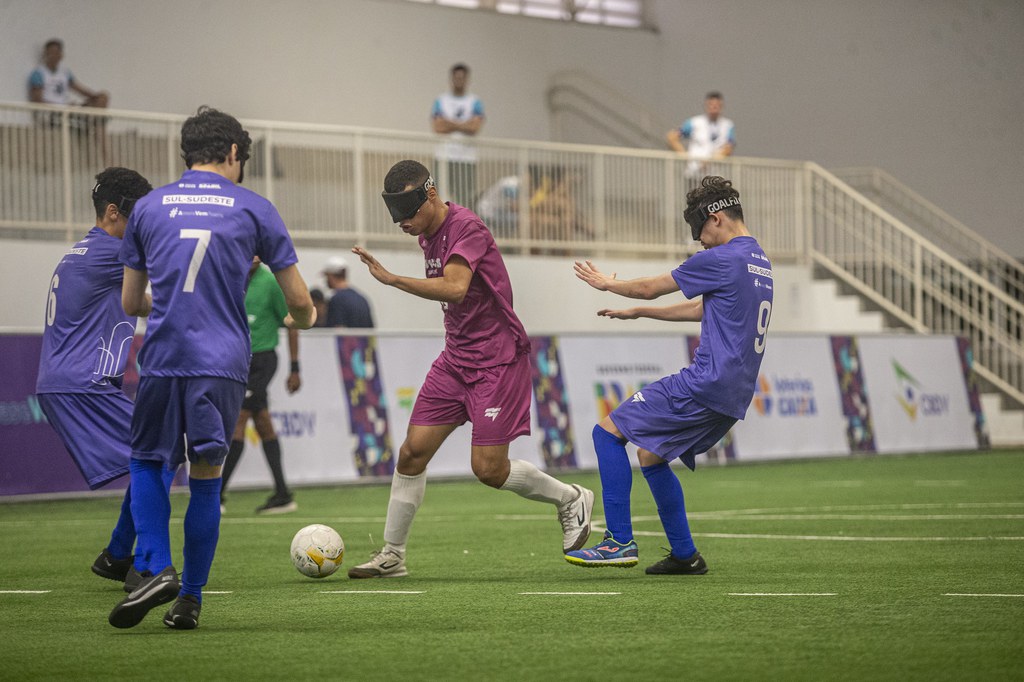 Raynã, atleta do time do Centro-Norte, que usa uniforme roxo com meião branco, domina a bola no campo de futebol de cegos e é marcado por três jogadores do Sul-Sudeste, que estão de azul. (Foto: Taba Benedicto/CBDV)